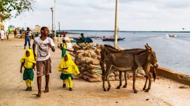 Lamu Town, Lamu Island, Kenya, Africa, Mar 12, 2018 A Kenyan teenager, accompanying two young Swahili girls in school uniform, along the dirt road next to the town’s waterfront - Copia