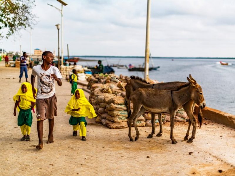 Lamu Town, Lamu Island, Kenya, Africa, Mar 12, 2018 A Kenyan teenager, accompanying two young Swahili girls in school uniform, along the dirt road next to the town’s waterfront - Copia