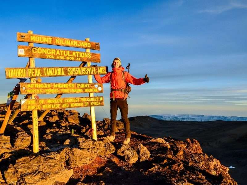 Kilimanjaro, Tanzania - March 11, 2015 A hiker standing on Uhuru Peak, the summit of Kibo and highest mountain in Africa, Mount Kilimanjaro at 5895m amsl. Summit sign and glacier in the background