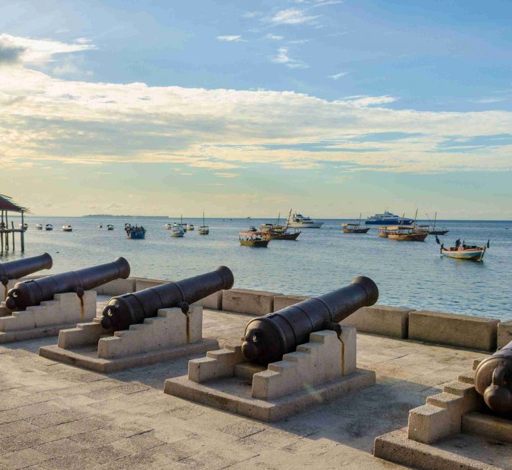 Five cannons lined up on the waterfront in Stone Town Zanzibar. Many fishing boats are out in the blue sea. It's a nice summer evening day with a few clouds in the sky and calm ocean
