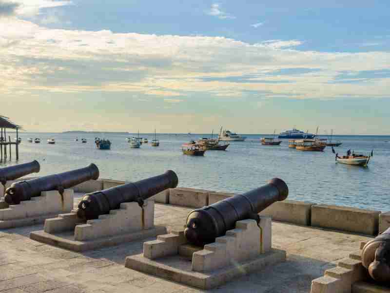 Five cannons lined up on the waterfront in Stone Town Zanzibar. Many fishing boats are out in the blue sea. It's a nice summer evening day with a few clouds in the sky and calm ocean